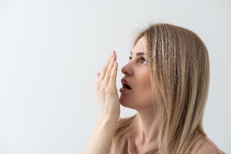 Eine junge Frau mit langen, blonden Haaren testet ihren Atem, indem sie ihre Hand vor ihren Mund hält. Sie schaut nach oben und trägt ein helles Oberteil. Der Hintergrund ist einfarbig weiß.